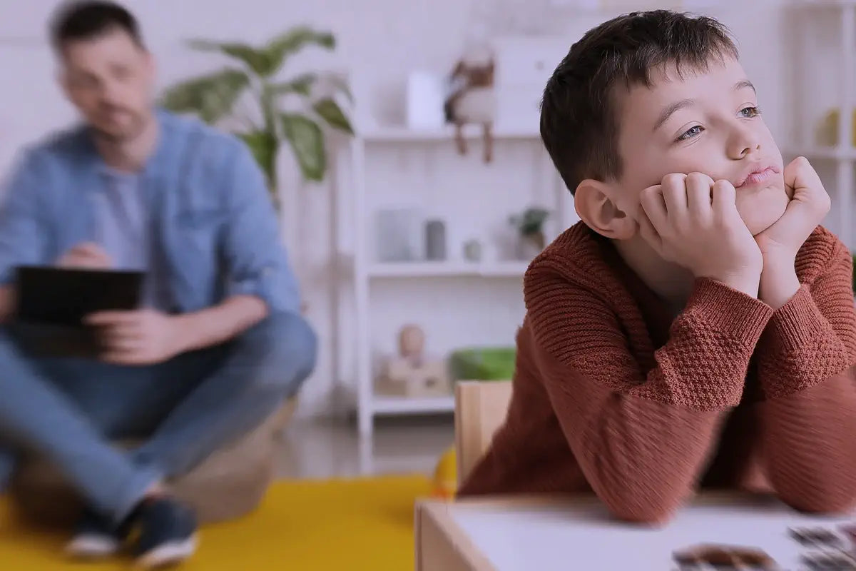 Bambino seduto a un tavolo con espressione sognante, mentre uno specialista lo osserva prendendo appunti in uno spazio educativo.