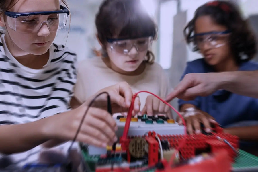 Bambine con occhiali protettivi lavorano insieme a un progetto STEM, assemblando componenti elettronici sotto la guida di un adulto.