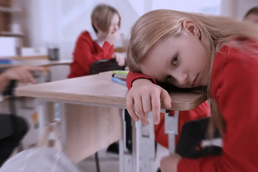 Bambina triste appoggiata al banco in classe, mentre alcuni compagni ridono e la indicano in sottofondo.