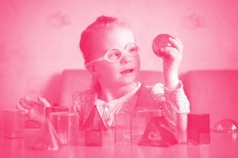 Bambina con disabilità intellettiva che osserva con curiosità una sfera trasparente, circondata da forme geometriche in plastica colorata